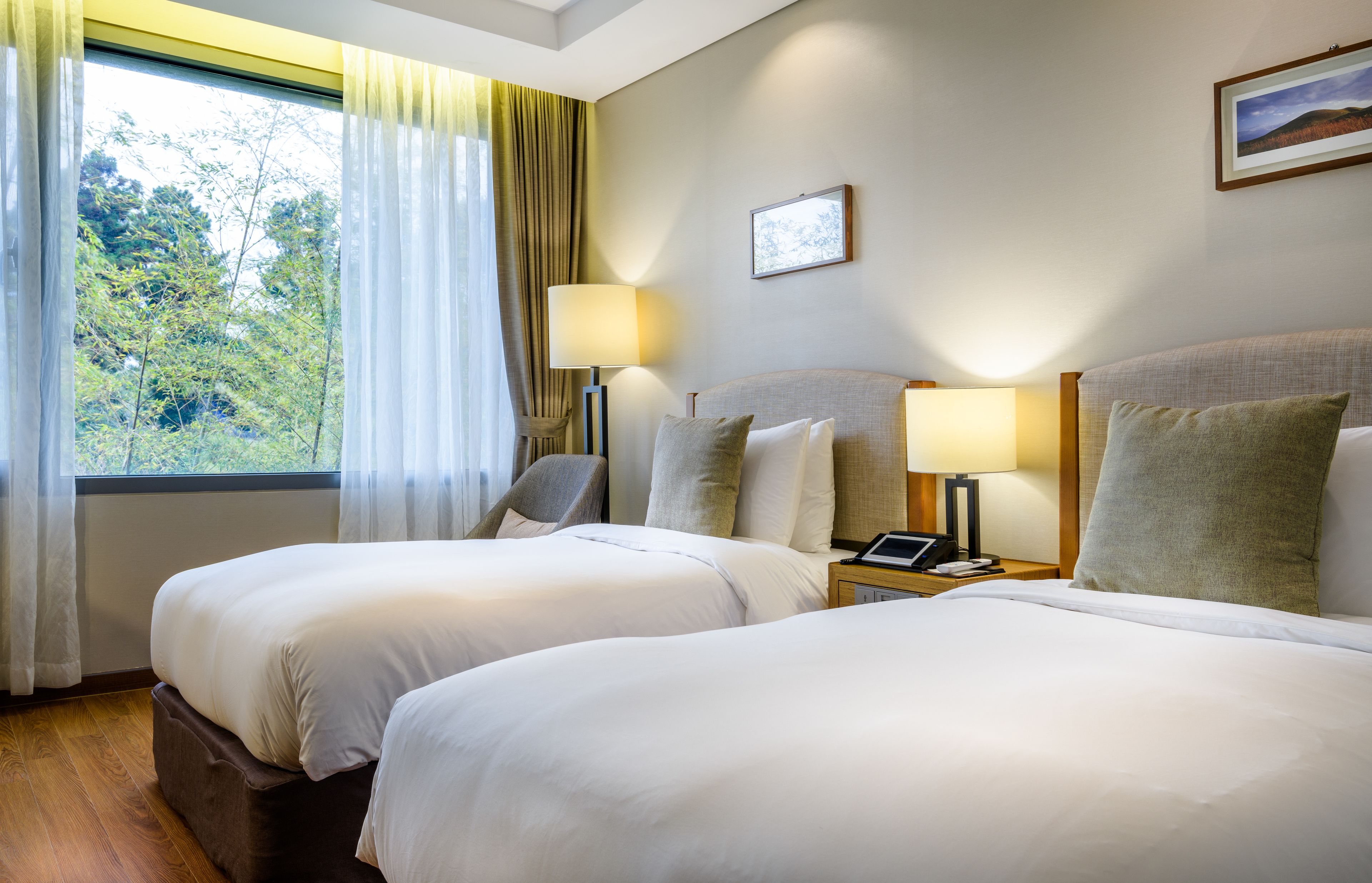 Une chambre d’hôtel avec deux lits, une fenêtre avec des rideaux, une chaise, des tables de chevet avec des lampes et un tableau encadré au mur.