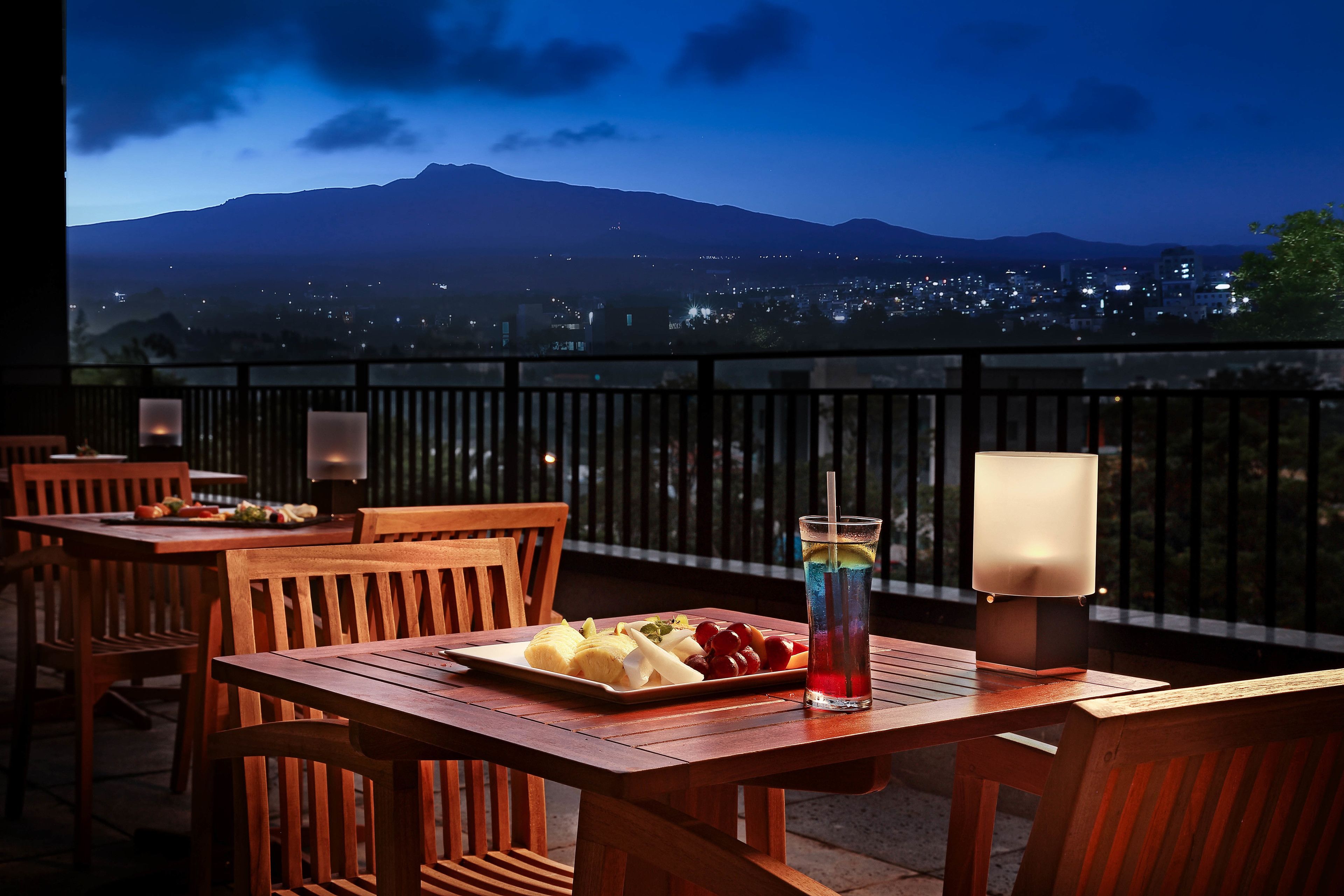 A wooden table with a plate of fruit and a drink, set against a balcony with a cityscape and mountains in the background.