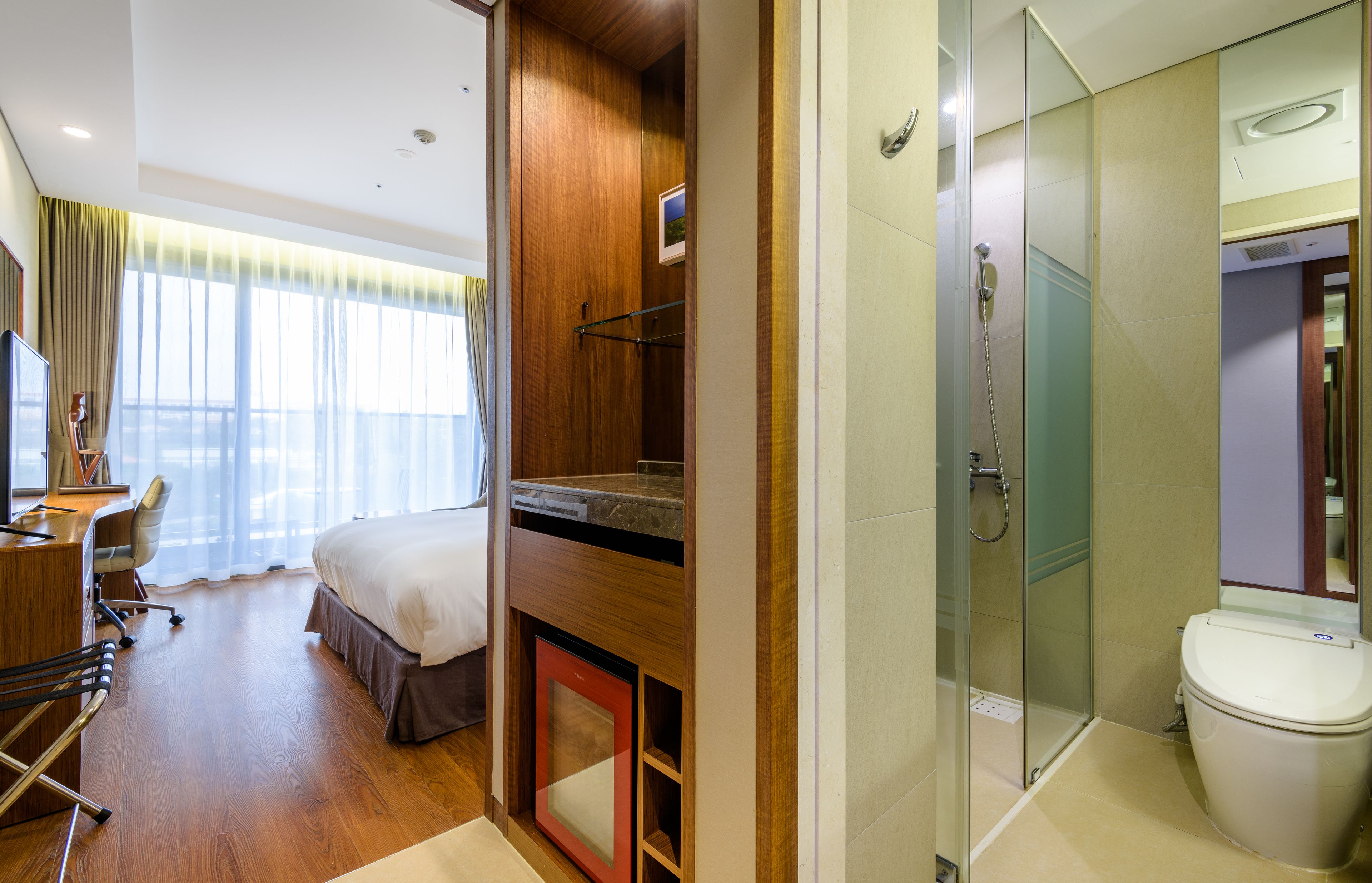 A hotel room with a bed, desk, chair, and a shower area with a glass door.