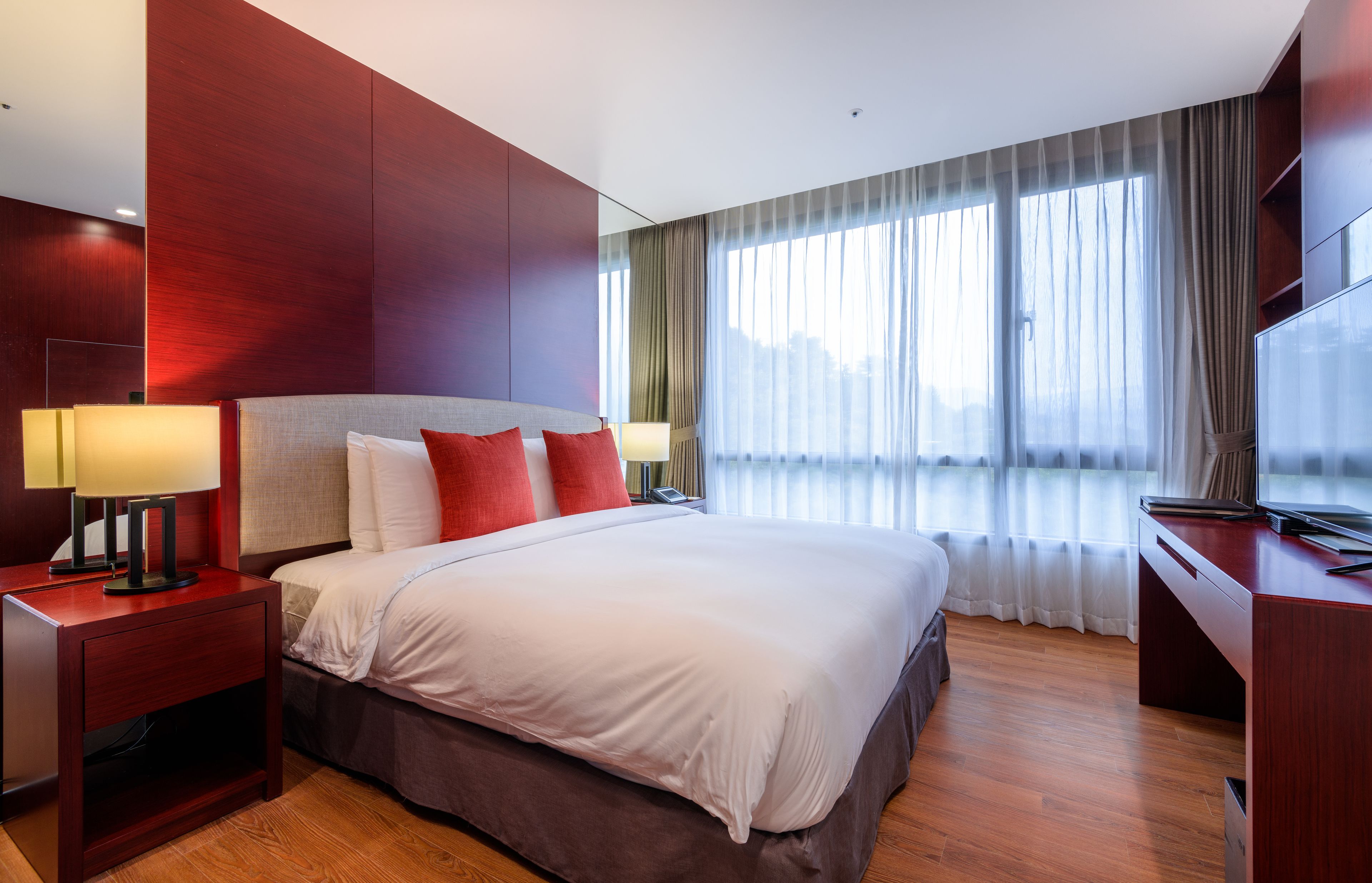 A modern hotel room with a large bed, two bedside tables with lamps, a desk with a flat-screen TV, and a large window with sheer curtains.
