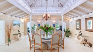 A spacious dining area with a high ceiling and natural light, featuring a central table with chairs, surrounded by potted plants and framed pictures on the walls. - Coconut Grove 1 by Island Villas (Westmoreland)