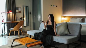 A modern hotel room with a grey sofa, a wooden side table, a desk with a red lamp, and a bed with white bedding. - THA City Loft by TH District (Bangkok)