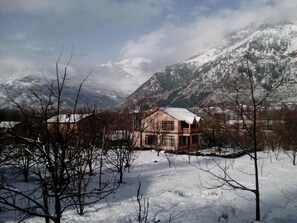 A house with a snow-covered roof, surrounded by snow-covered ground and mountains in the background.