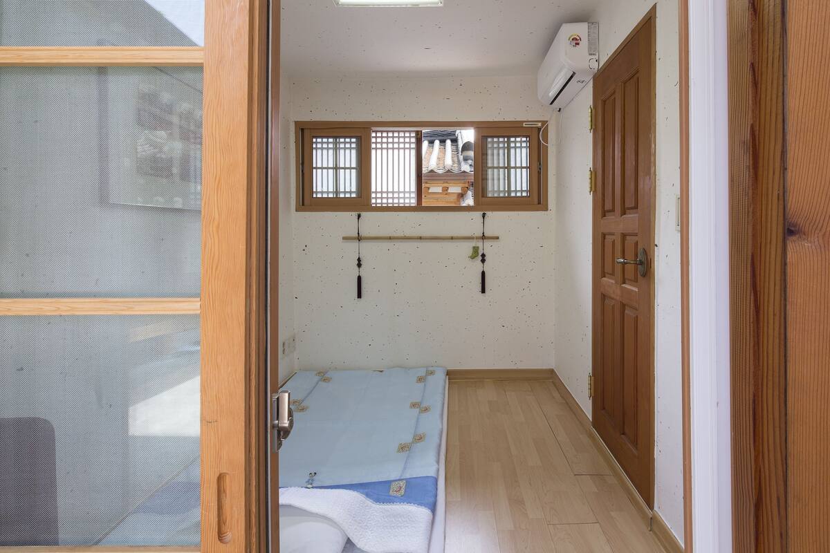 A small room with a bed, a window with a wooden frame, a wooden door, and a wall-mounted air conditioner.