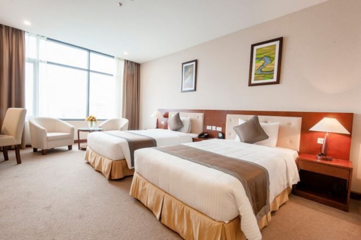 A hotel room with two beds, a sitting area with a chair, a small table, and a nightstand with a lamp.