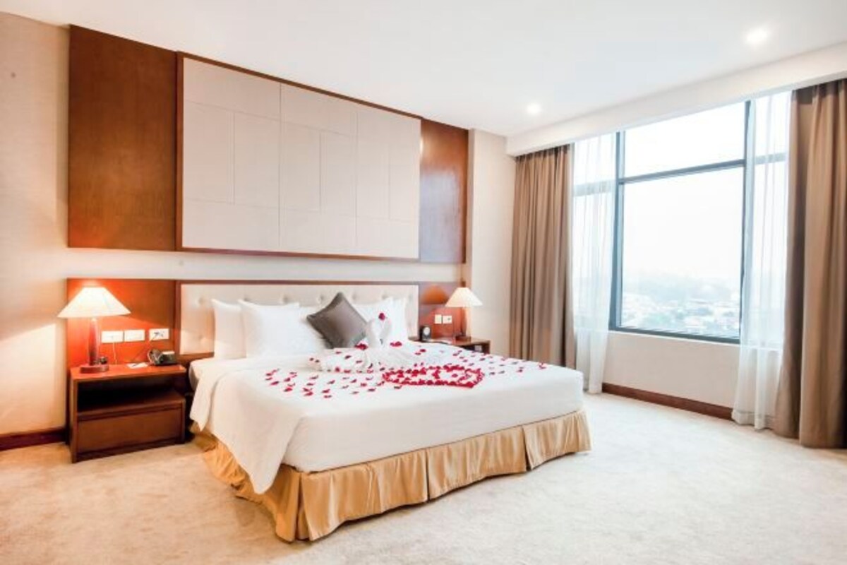 A hotel room with a large bed, two bedside tables with lamps, a large window with curtains, and a city view.