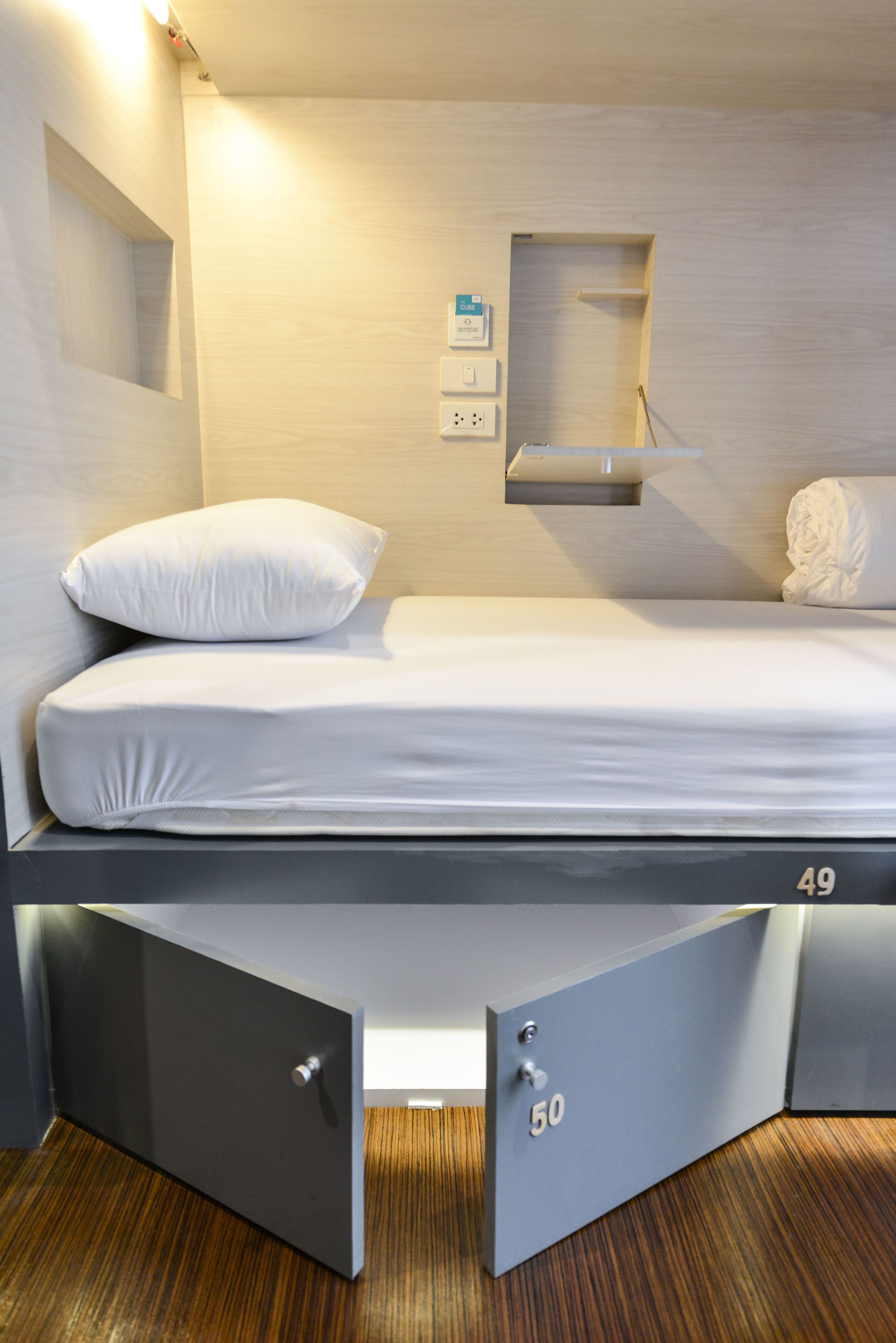 A modern hotel room with a single bed, a small bedside table, a wall-mounted power outlet, and a built-in shelf.