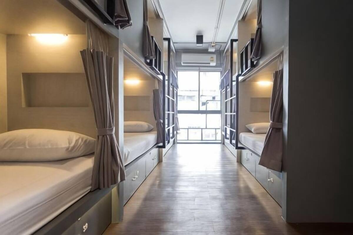 A narrow corridor with bunk beds on either side, a window at the end, and a door leading to a larger room.