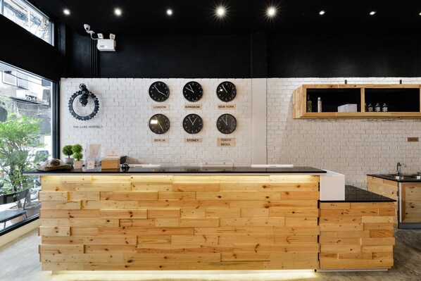 A modern kitchen with a wooden counter, a white brick wall, and clocks displaying times from various cities. - The Cube Hostel Silom (Bangkok)