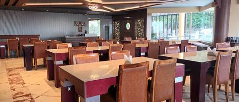 Un restaurant spacieux avec de hauts plafonds, des poutres en bois et un sol en marbre. Les tables et les chaises sont disposées pour accueillir les clients.