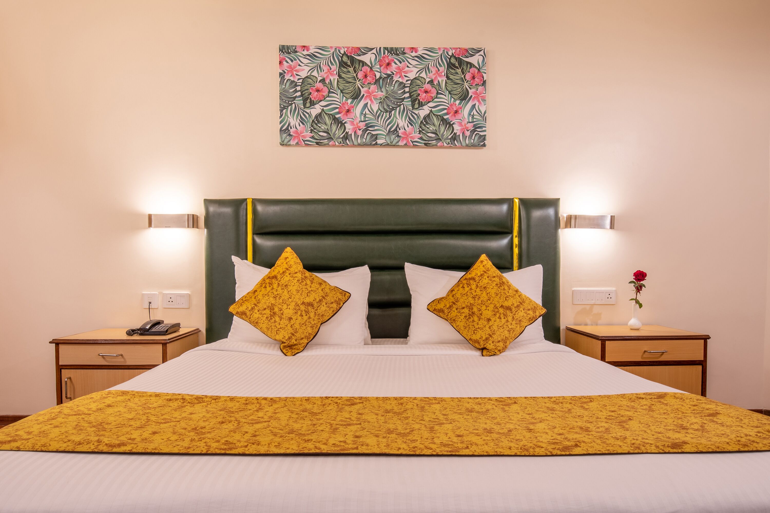 Une chambre d’hôtel avec un grand lit, deux tables de chevet, une petite table, une œuvre d’art florale au-dessus du lit et un vase contenant une seule rose rouge sur la petite table.