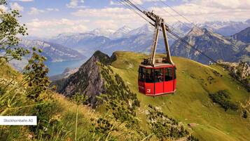 A red cable car with the number 3, suspended by cables, traveling over a mountainous landscape with a lake and snow-capped peaks in the background.
