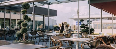 Un café al aire libre con mesas y sillas, una escultura de árbol alta y un edificio moderno al fondo.