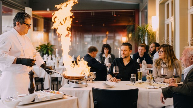 Un chef prépare un plat flambé à l’aide d’une poêle sur une table dans un restaurant.