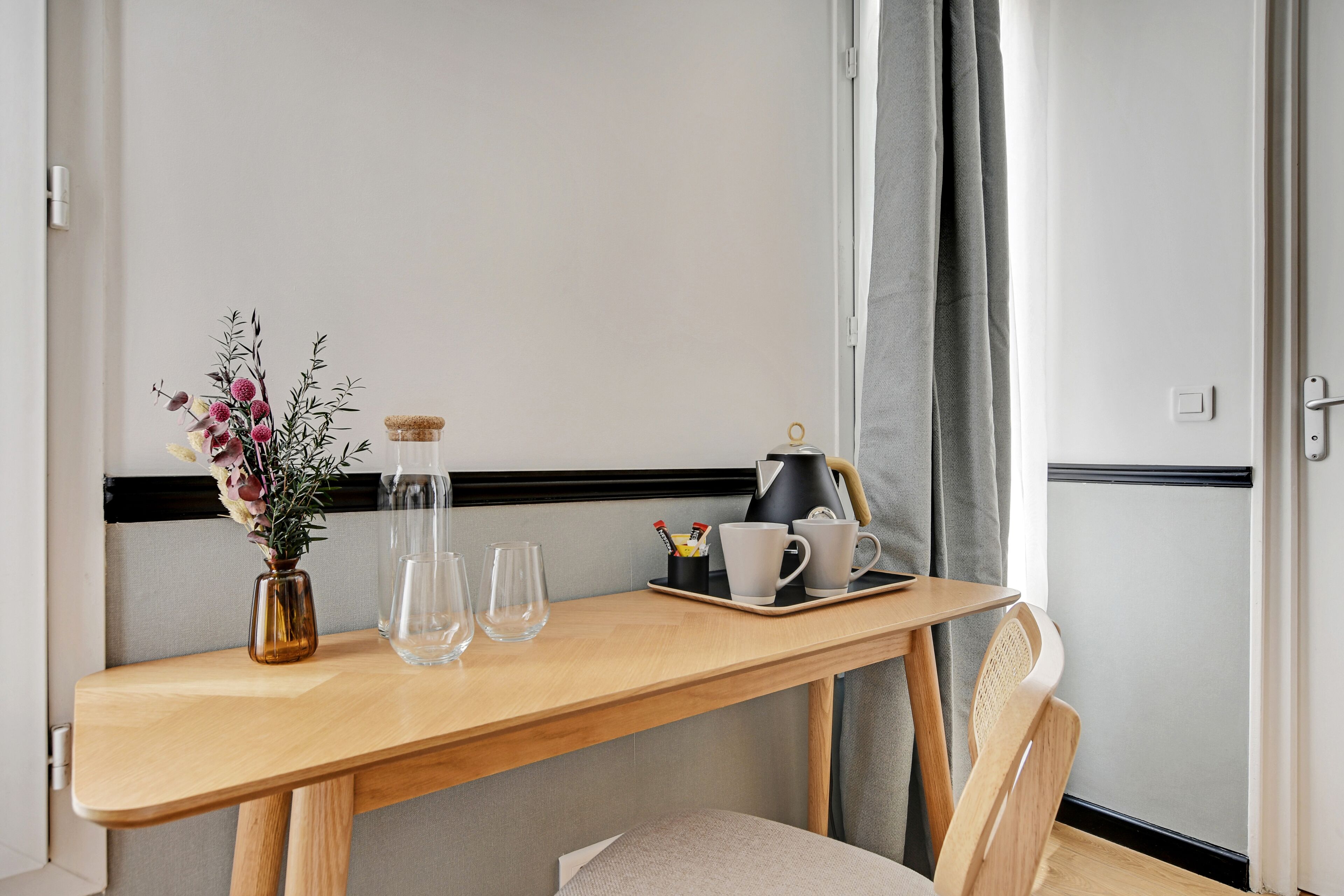 Une table en bois sur laquelle se trouvent un vase de fleurs, deux verres, une cafetière et un plateau avec des tasses et un petit récipient.