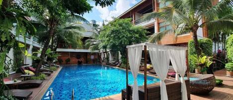 A pool area with a wooden deck, a covered seating area, and lush greenery.