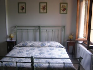 A bedroom with a metal bed frame, floral bedding, a small bedside table with a lamp, and a window with a view of the outside.