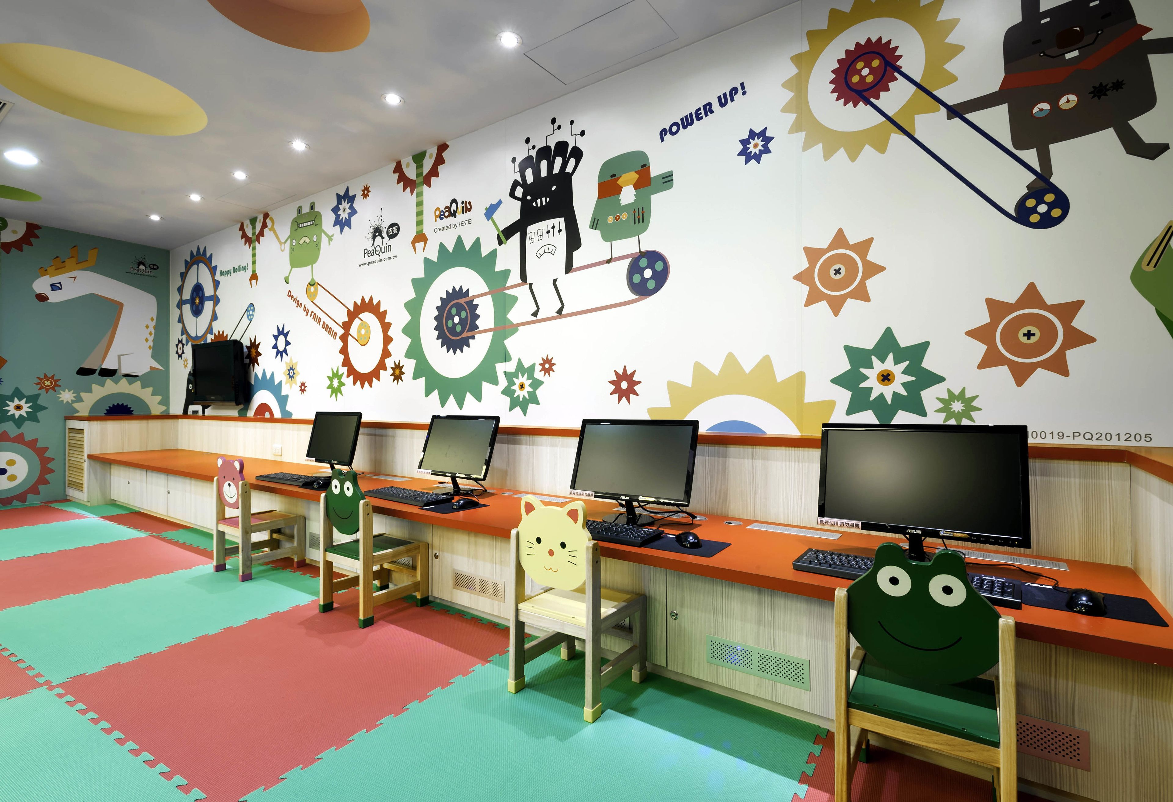 Um laboratório de informática infantil com várias estações de trabalho, cada uma com monitor, teclado e rato. O espaço possui decorações coloridas nas paredes e um tapete vermelho e verde no chão.
