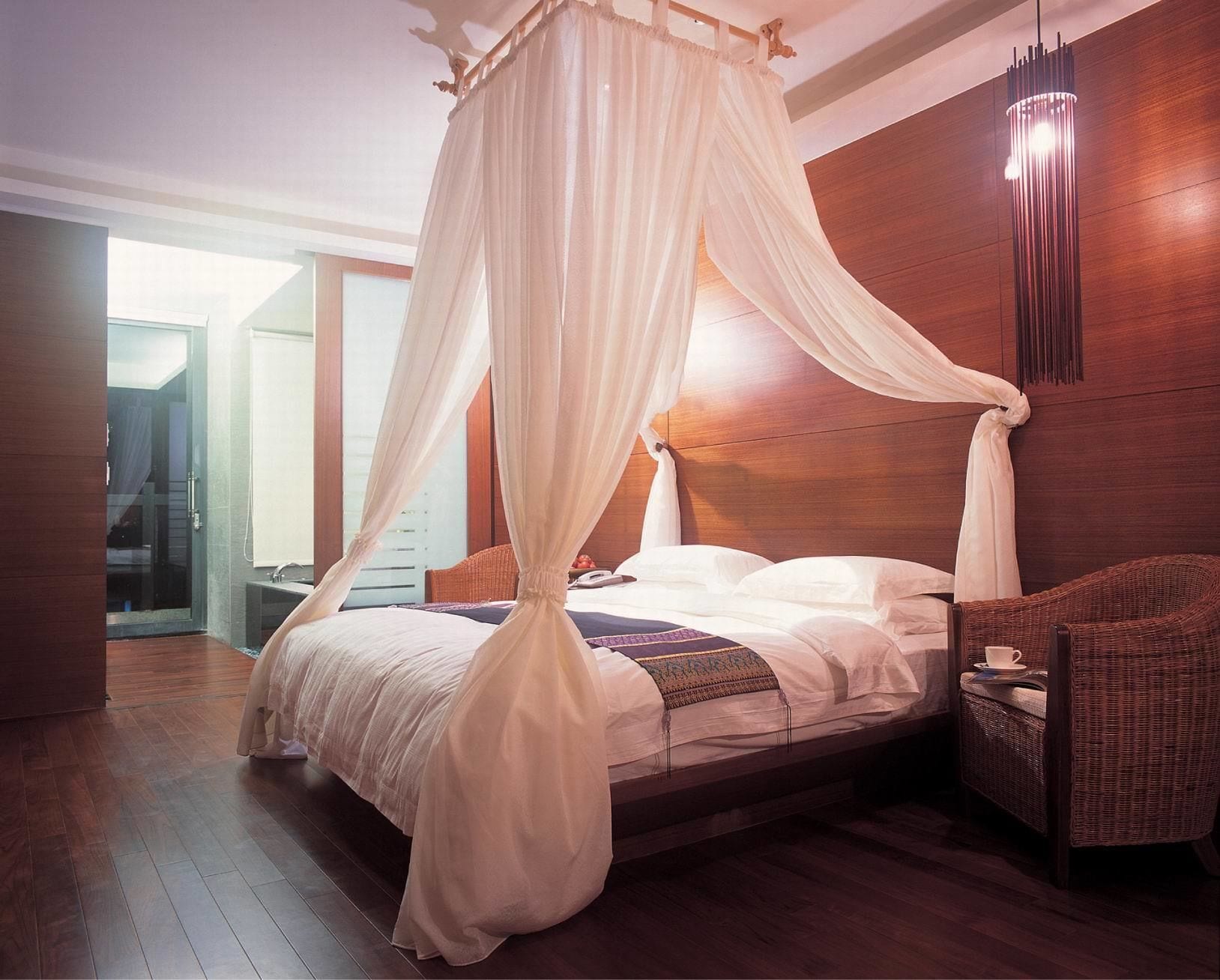A bedroom with a four-poster canopy bed, a wooden headboard, a wicker side table, and a view of the outside.