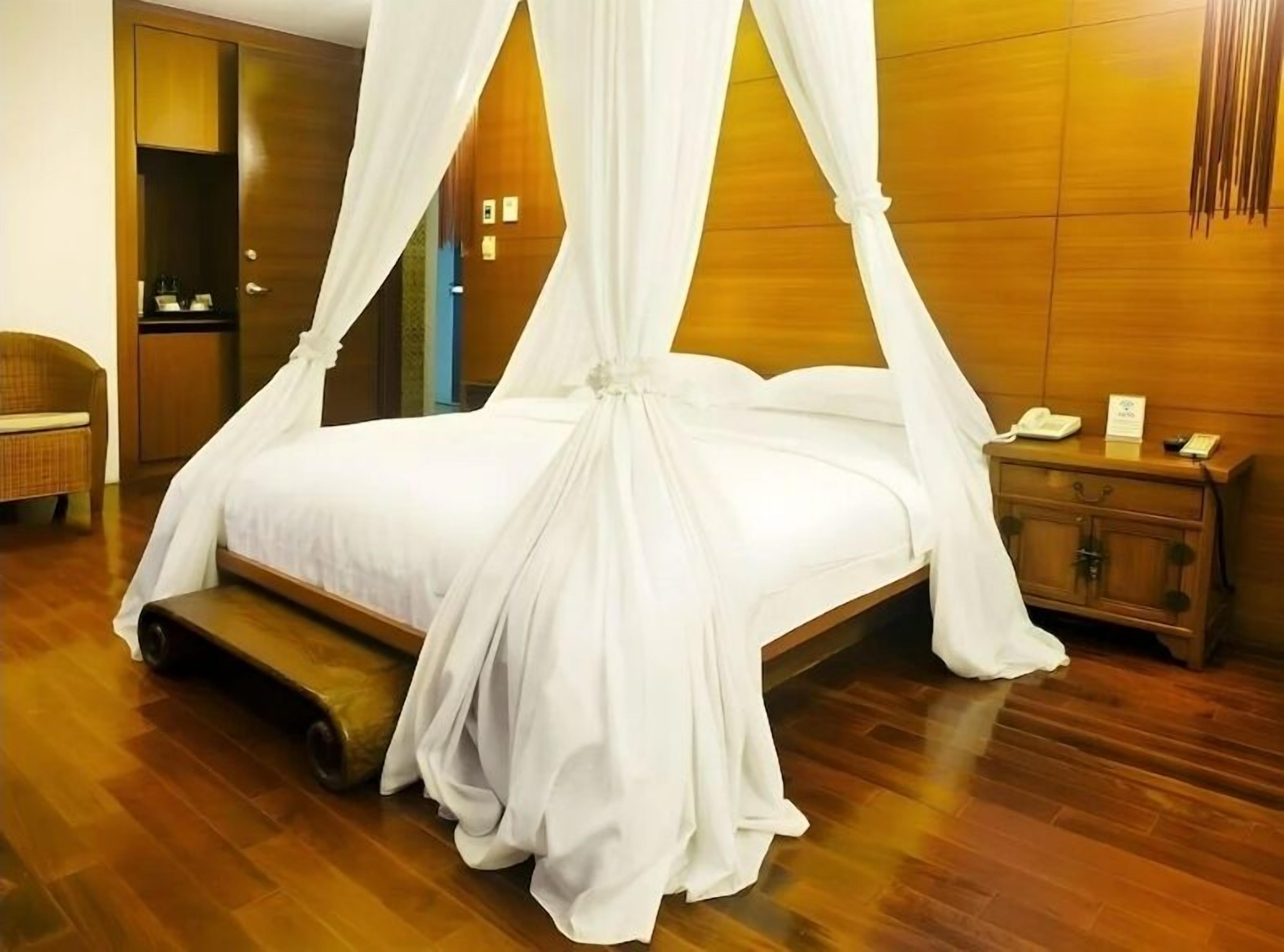 Una cama con dosel, ropa de cama blanca y un dosel, una mesita de noche de madera con un teléfono, y suelo de madera.