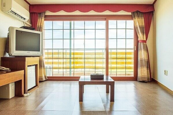 A room with a television on a wooden stand, a small table, and a sliding glass door leading to a balcony with yellow railing. - LK Hotel (Jeju City)