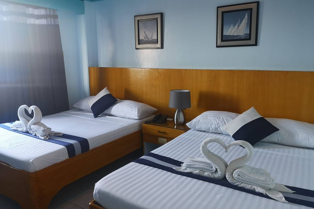 Two beds with white linens, a wooden headboard, a bedside table with a lamp, and a curtain.