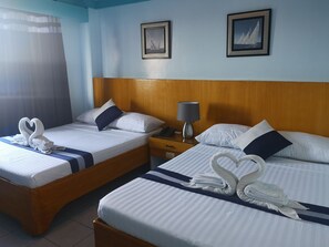 Duas camas com roupa de cama branca, uma cabeceira de madeira, uma mesa de cabeceira com abajur e uma cortina.