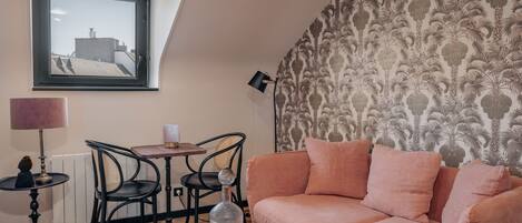 Una sala de estar acogedora con un sofá rosa, una mesita y una silla. Hay una ventana con vistas a edificios, un papel tapiz estampado y una lámpara sobre la mesita.