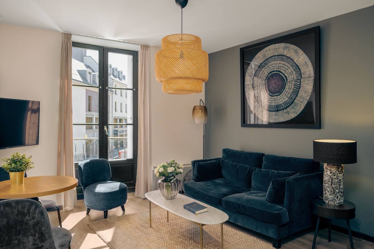 A modern living room with a blue sofa, a round coffee table, and a large window with a view of buildings.