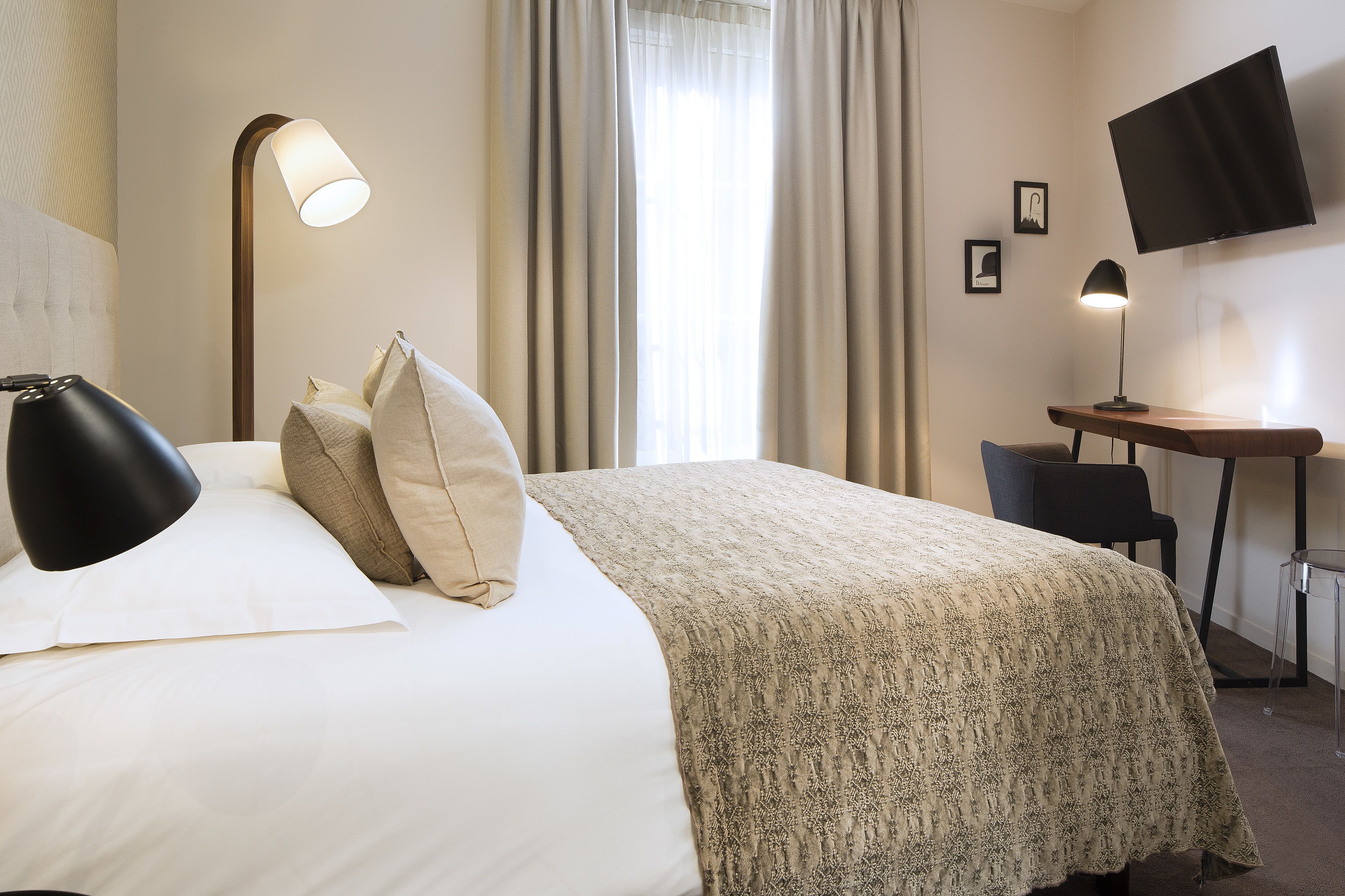 A hotel room with a bed, two bedside lamps, a desk with a chair, a flat-screen TV, and curtains.