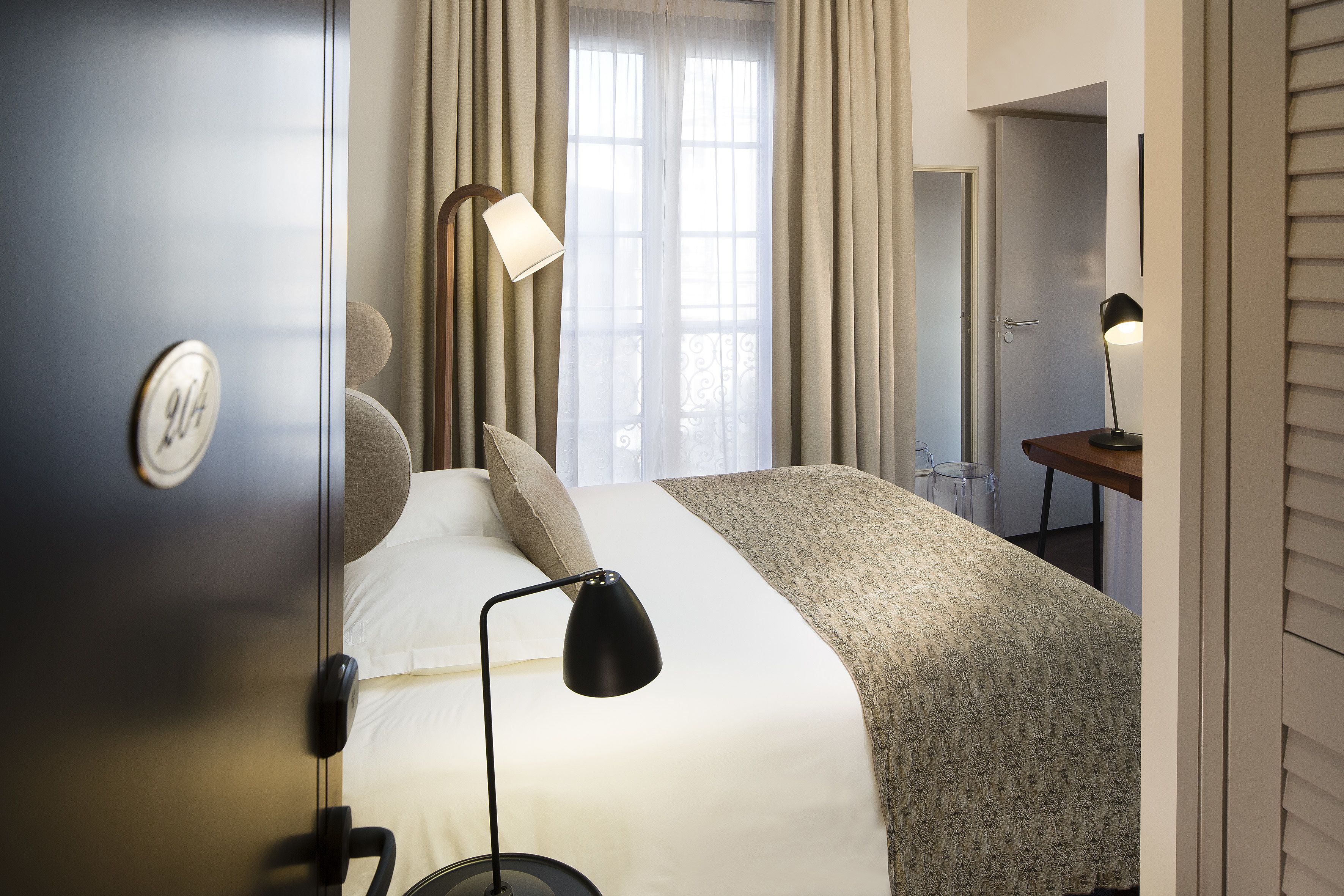 A hotel room with a bed, a desk, a lamp, and a window with curtains.