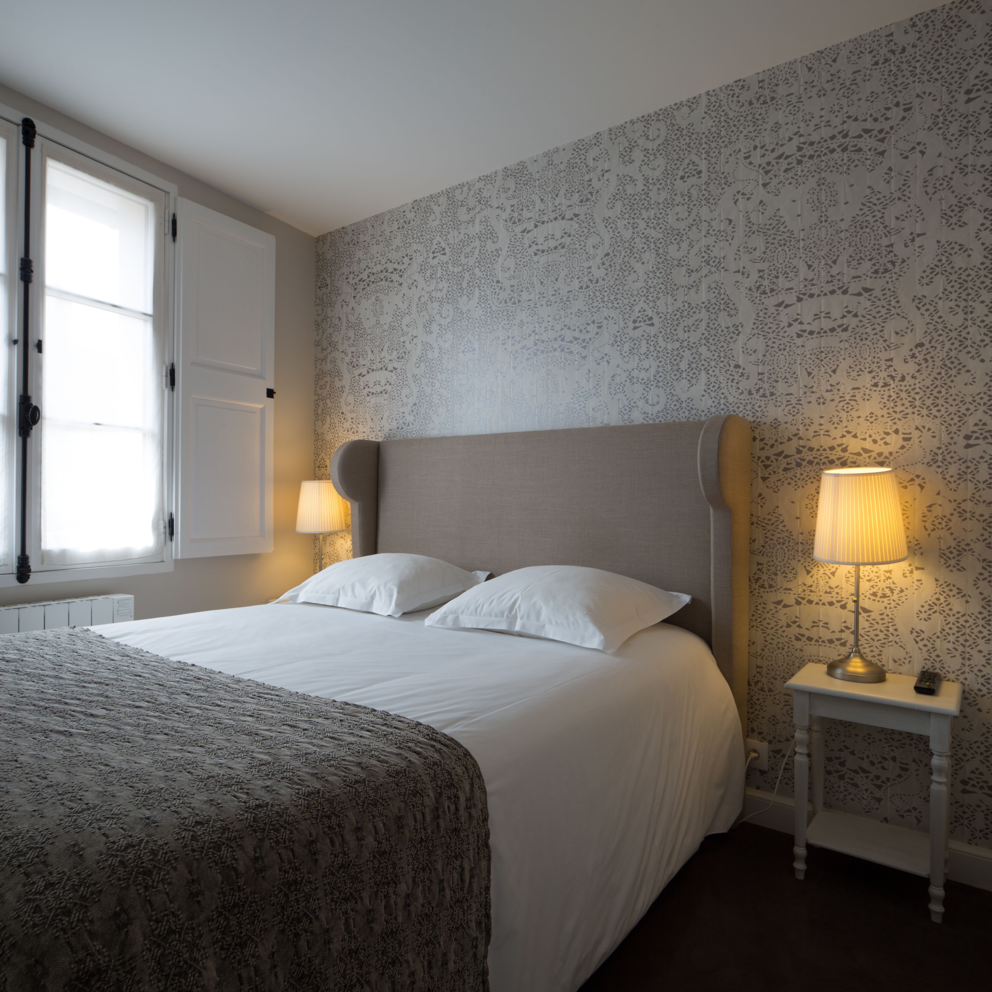 Un lit bien fait, avec du linge de lit blanc et deux oreillers, une table de chevet avec une lampe, et une fenêtre donnant sur l’extérieur.