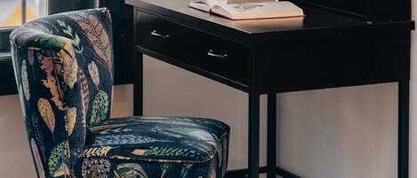 A black desk with a book and a small potted plant. A chair with a leaf pattern is in front of the desk. A framed picture hangs on the wall.