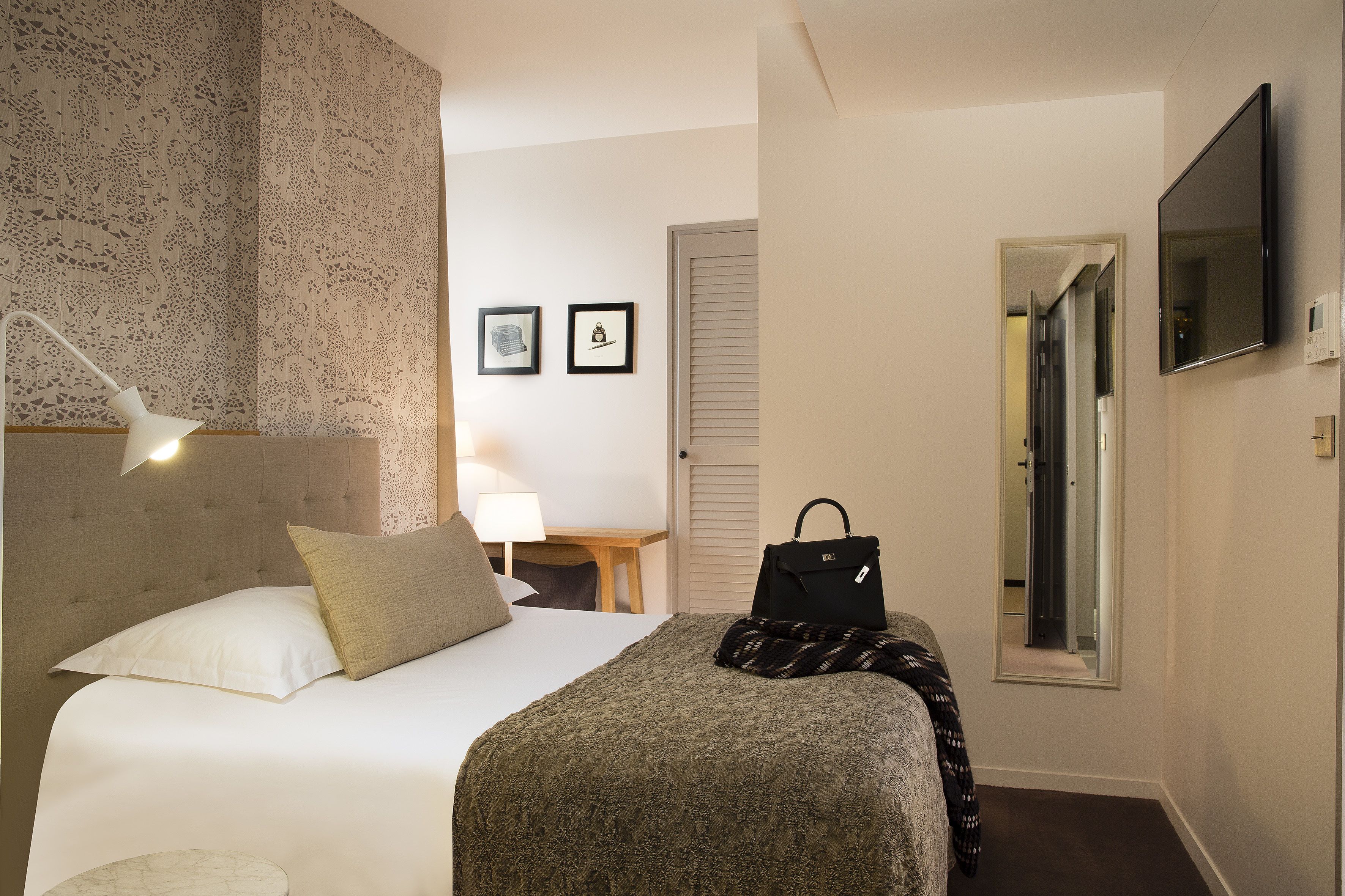 A hotel room with a bed, a bedside lamp, a nightstand, a mirror, and a door leading to another room.