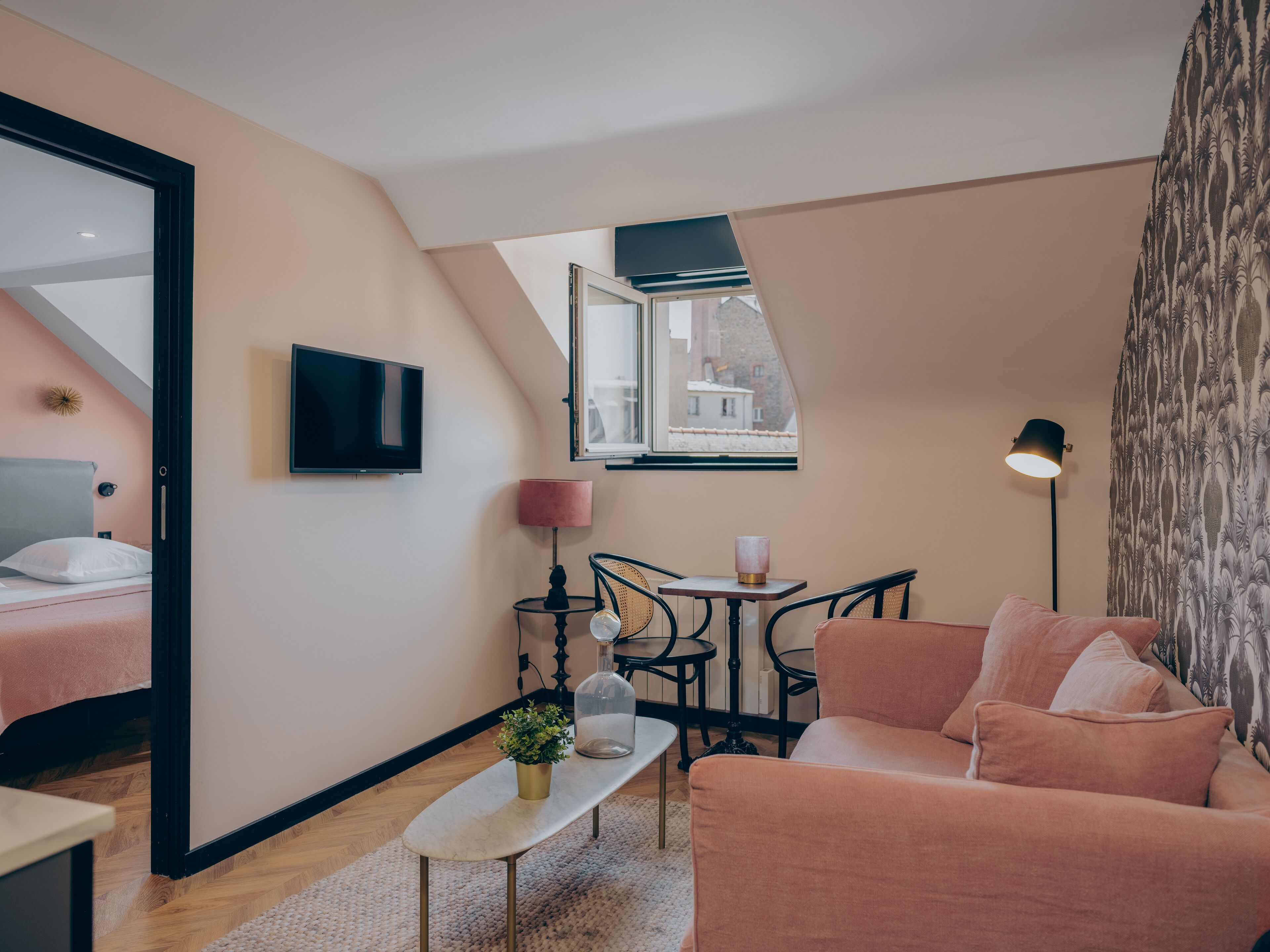 Une pièce confortable avec un lit, un canapé, une petite table agrémentée d’une plante, une télévision, une lampe et une fenêtre donnant sur des immeubles.