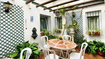 Un patio con una mesa redonda y sillas blancas, rodeado de plantas en macetas y una pared con celosía.