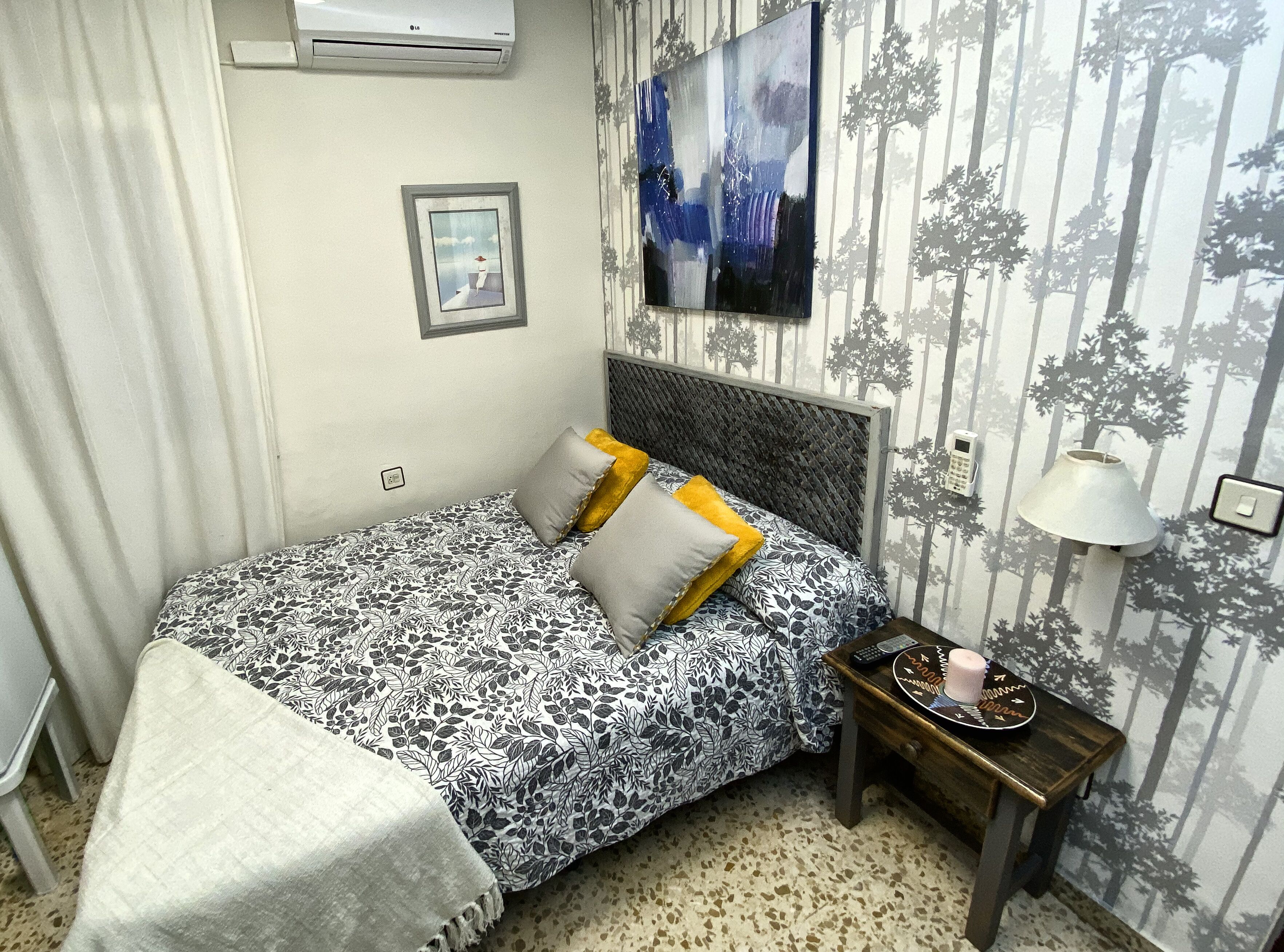 Un dormitorio ordenado con una colcha estampada, almohadas, una mesita de noche con una lámpara y un aire acondicionado de pared.