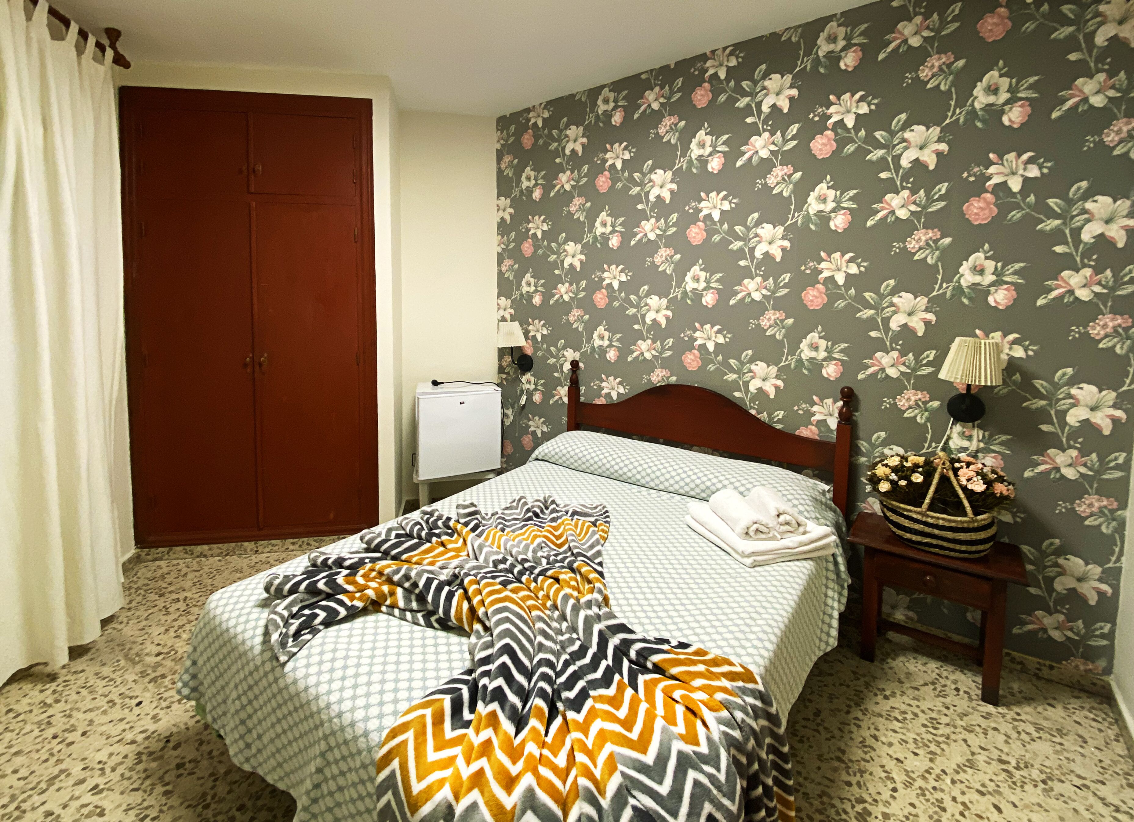 A bedroom with a bed, a nightstand, a lamp, a floral wallpaper, and a wardrobe.
