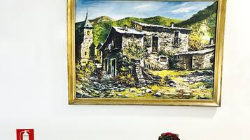 A painting of a rural landscape with a stone building and a person standing in the foreground. Below the painting, there is a wooden table with a vase of flowers, a sign with a price, and a fire extinguisher on the wall.