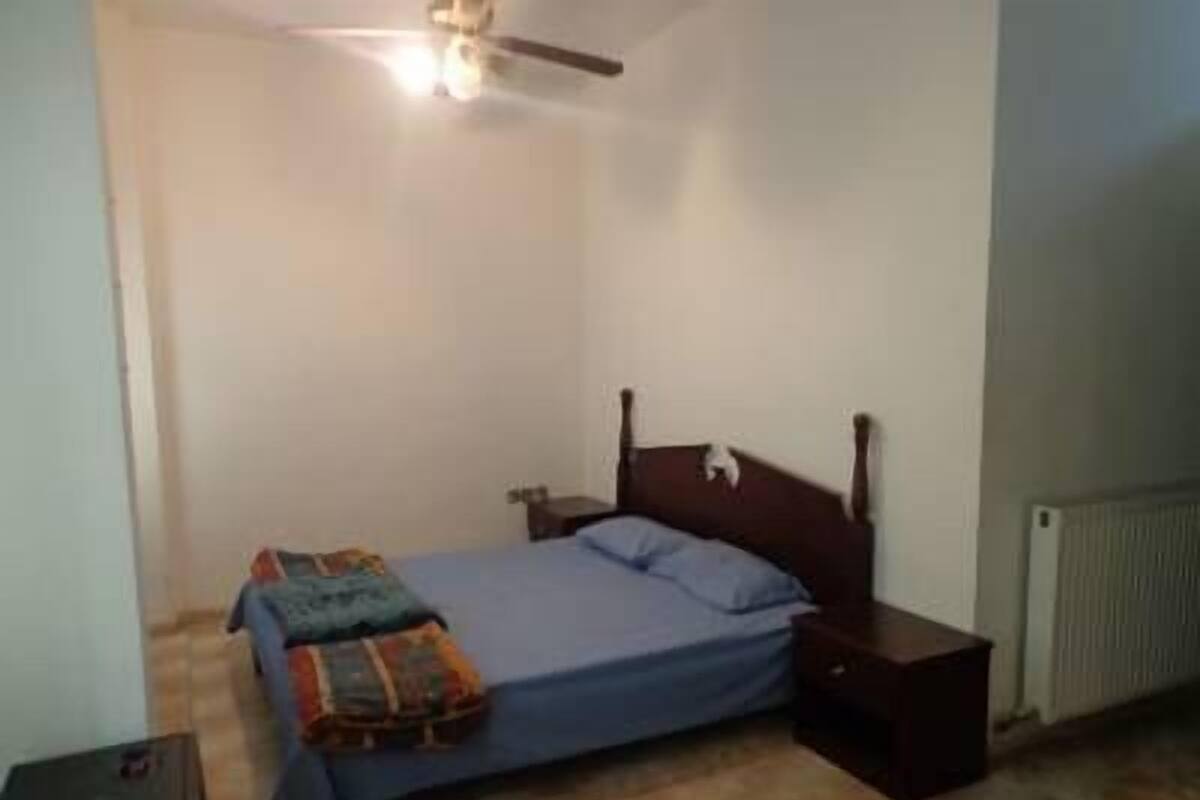 Een kleine, eenvoudige slaapkamer met een enkel bed, een nachtkastje, een plafondventilator en een radiator.