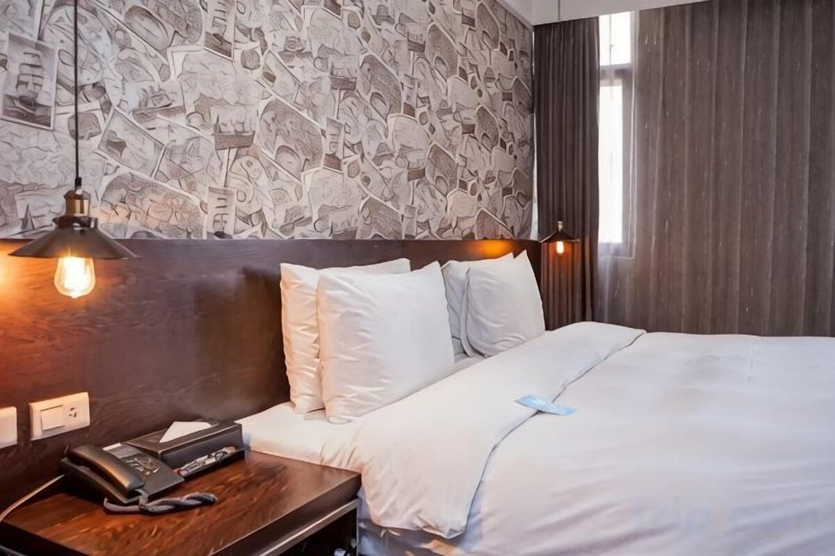 A hotel room with a patterned wallpaper, a wooden headboard, a bedside table with a phone, and a bed with white bedding.