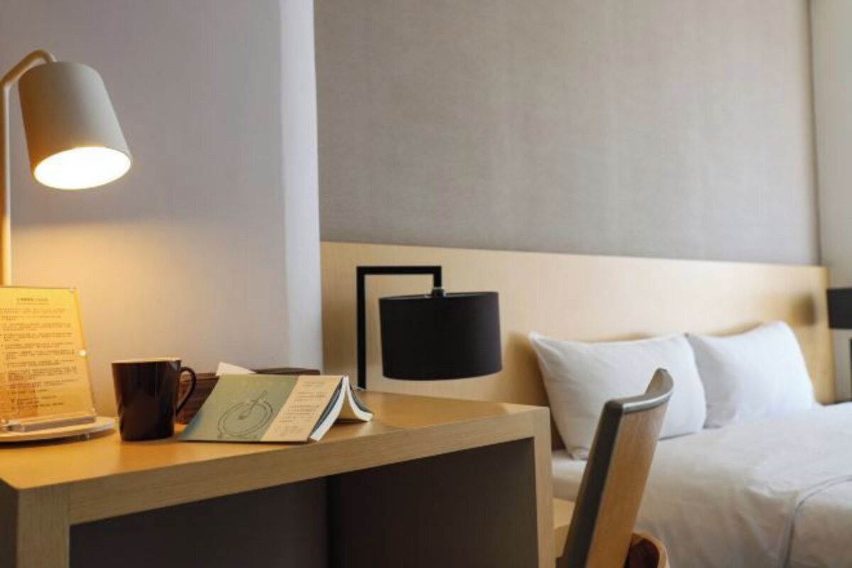 Habitación de hotel con un escritorio de madera, una lámpara de escritorio negra, una silla negra, una cama con ropa blanca y una lámpara negra de pared.