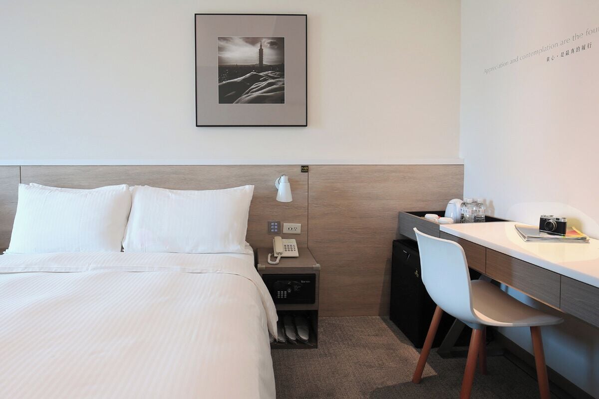 A hotel room with a bed, a desk, a chair, a phone, a small cabinet, and a framed picture on the wall.