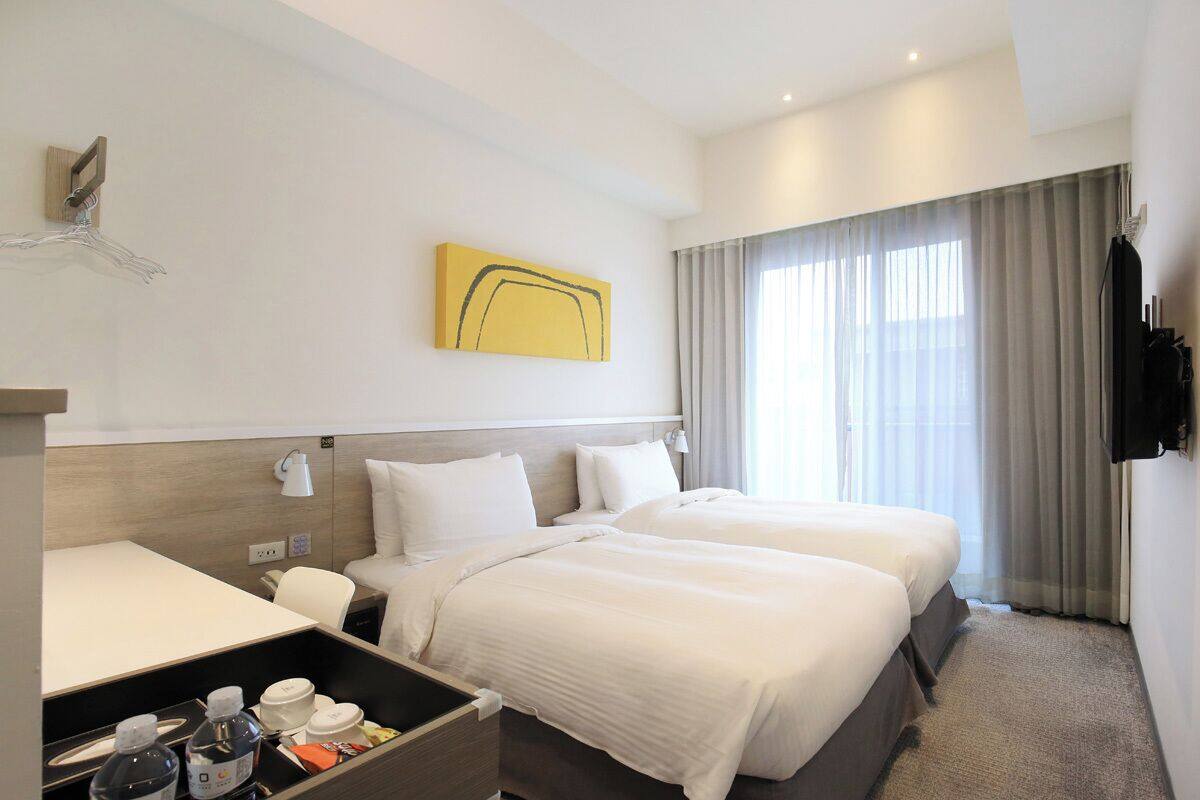 A hotel room with two beds, a wooden headboard, a bedside table, a lamp, a TV, and a window with curtains.