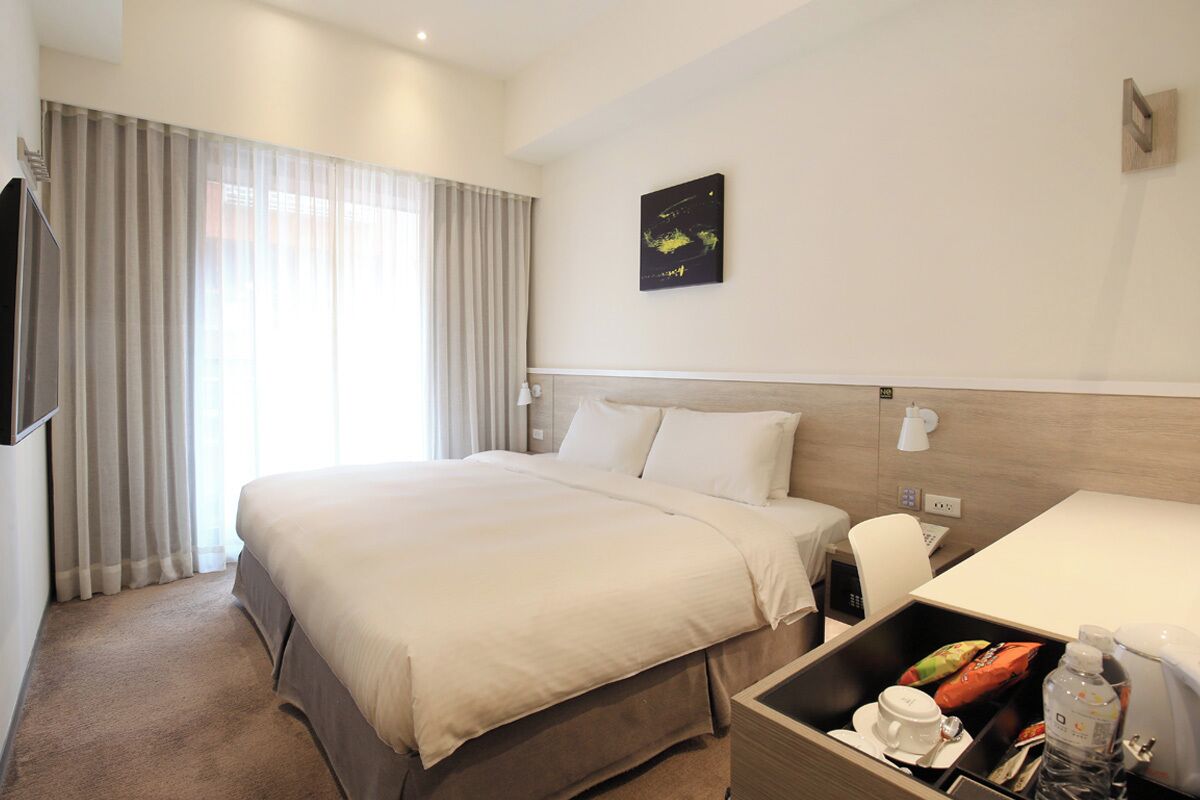 A hotel room with a large bed, a desk, a chair, a TV, a wall-mounted artwork, and a window with curtains.
