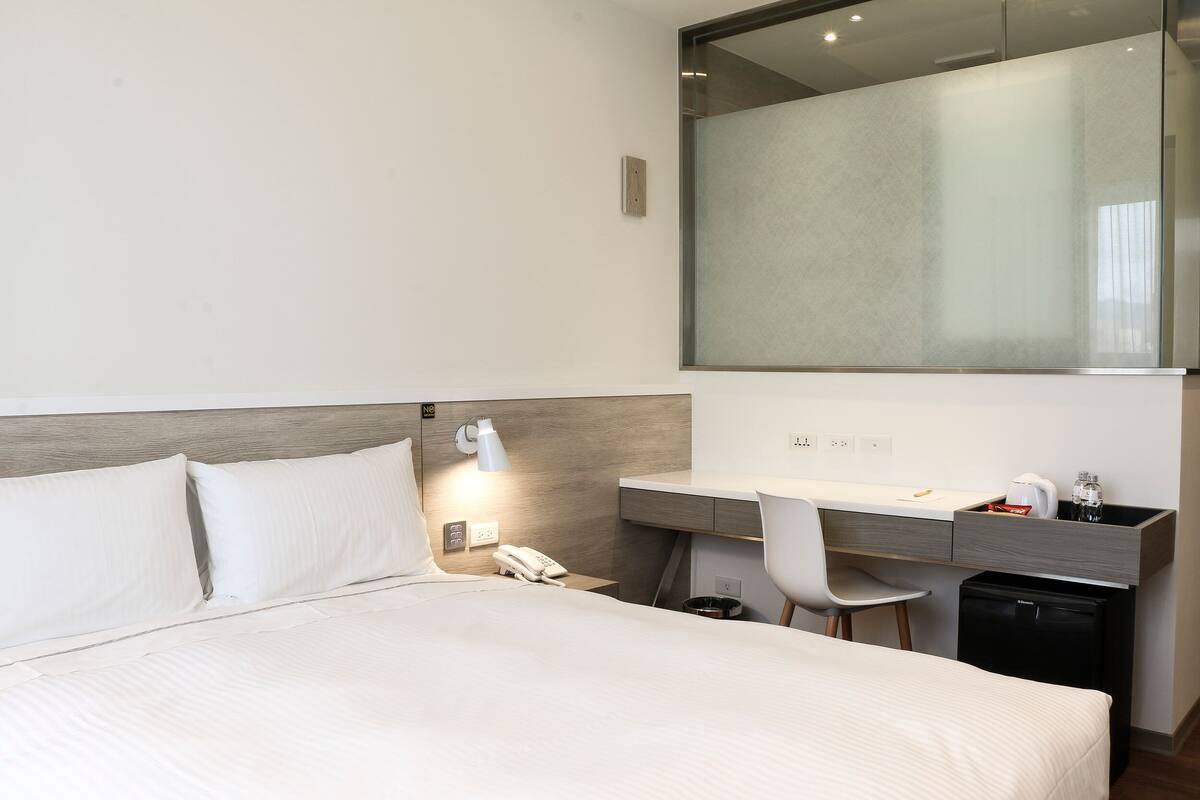 A hotel room with a bed, a desk with a chair, a small refrigerator, a phone, and a mirror.