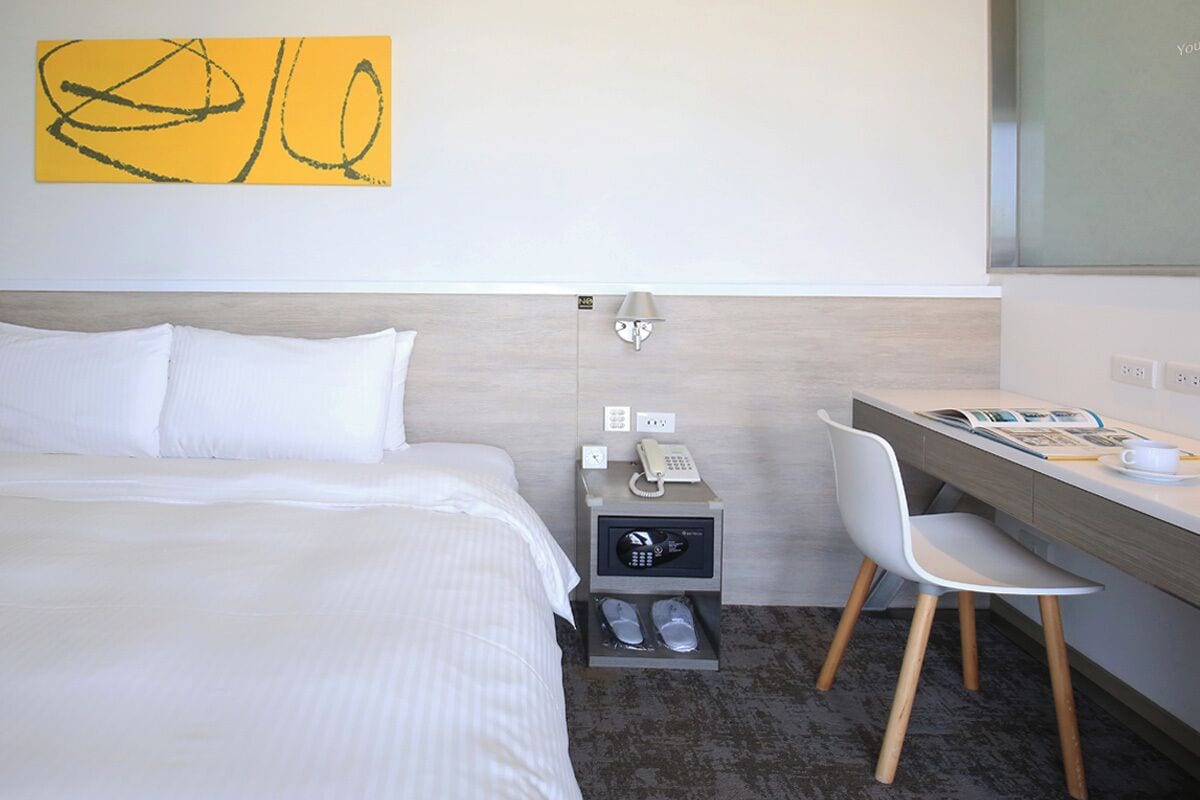 Quarto de hotel com cama, secretária, cadeira, telefone e um móvel para guardar sapatos.