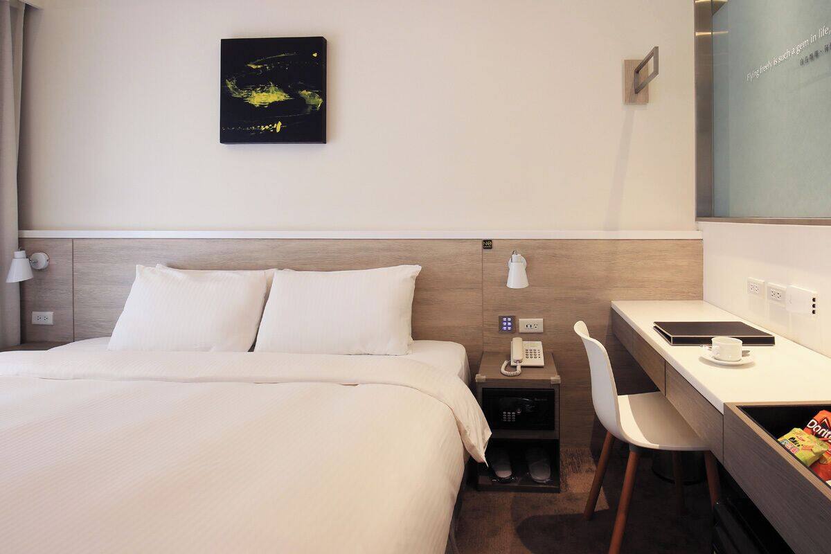 A hotel room with a bed, a desk, a chair, a phone, a lamp, and a wall-mounted artwork.