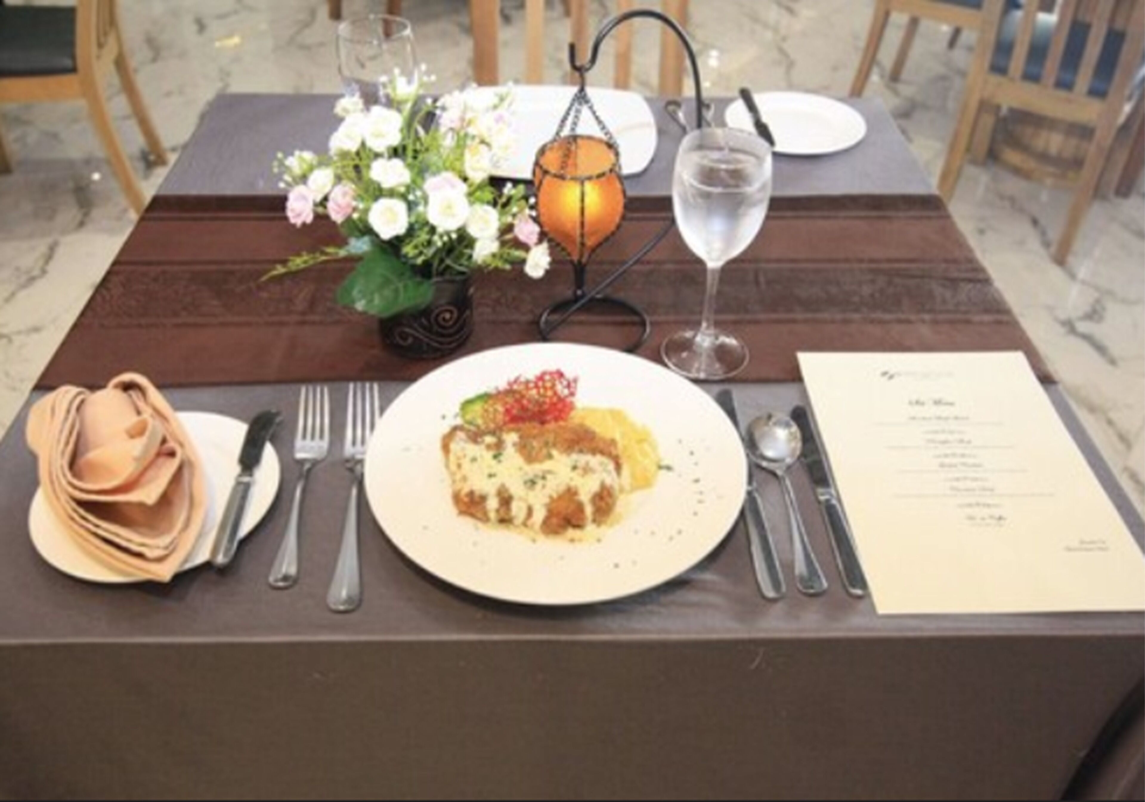 A dining table set with a plate of food, cutlery, a glass of water, a menu, and a small flower arrangement.