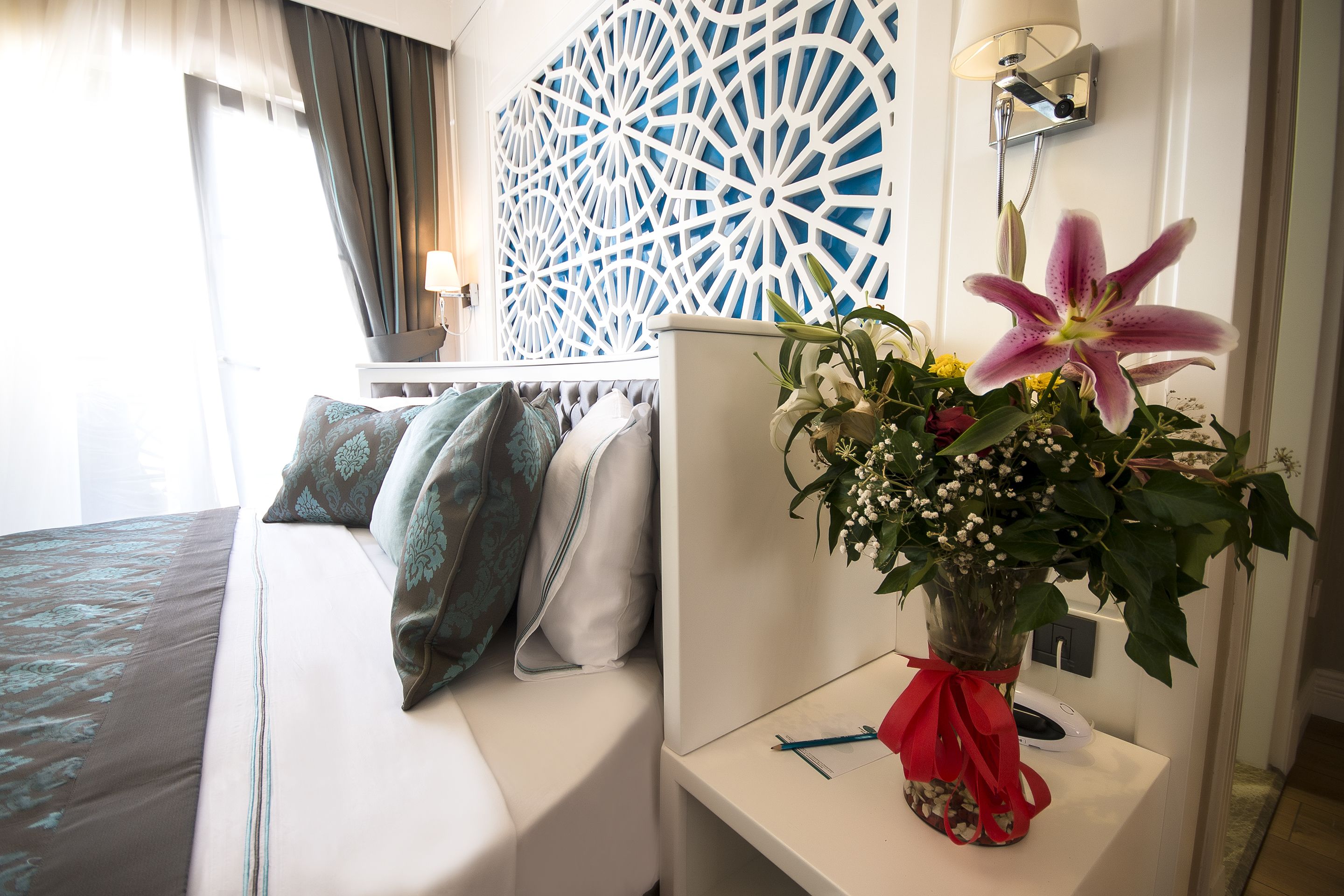 A hotel room with a bed, a bedside table with a vase of flowers, a decorative wall panel, and a lamp.